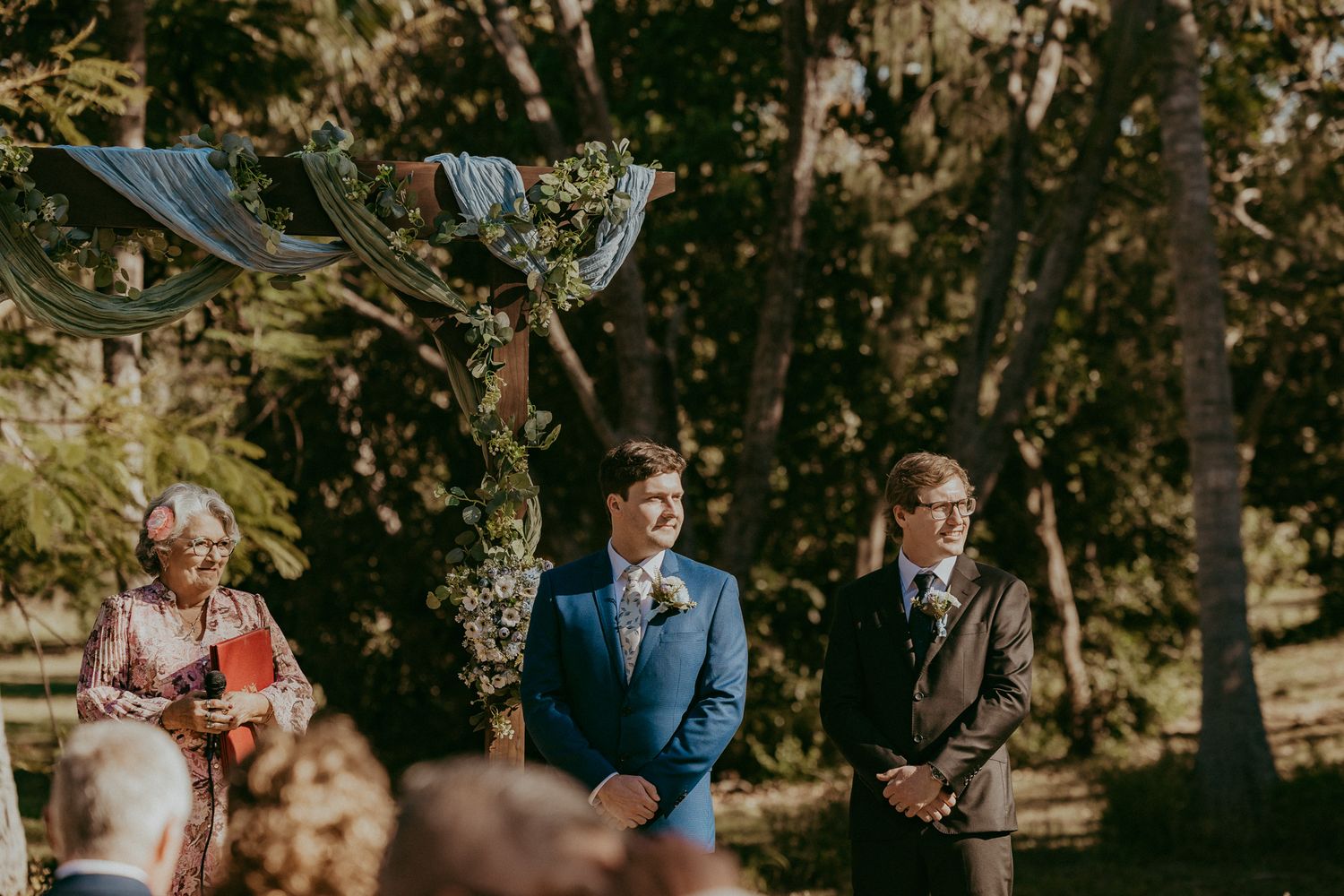 Groom stands at the altar beneath a decorated wedding arch during an outdoor ceremony.