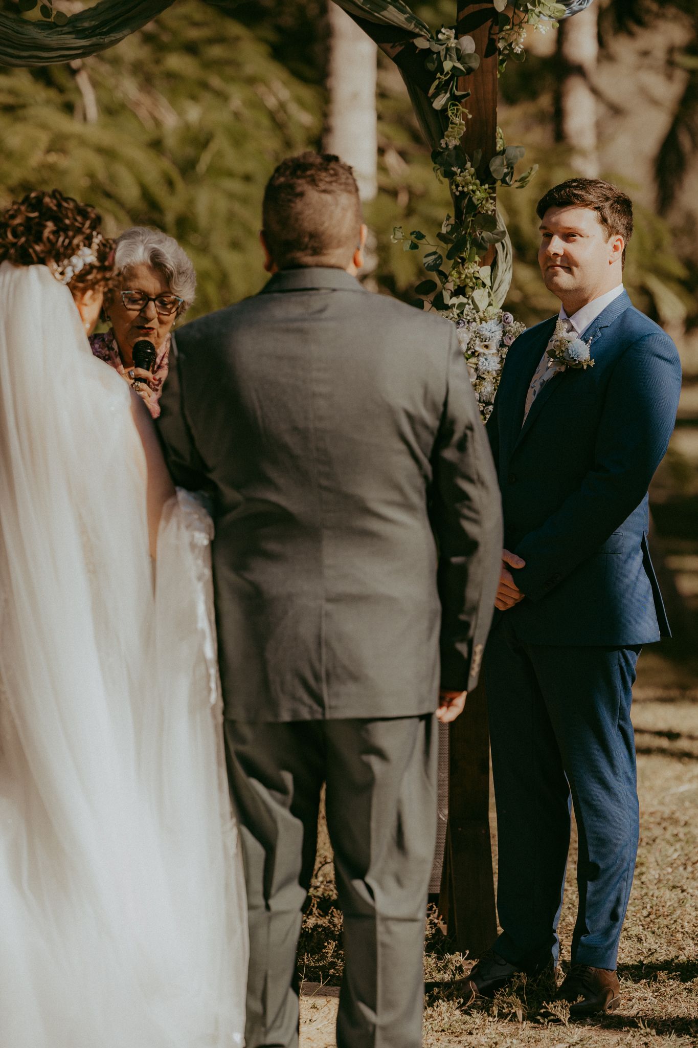 Groom in blue suit smiles during outdoor wedding ceremony beneath a floral decorated arch.