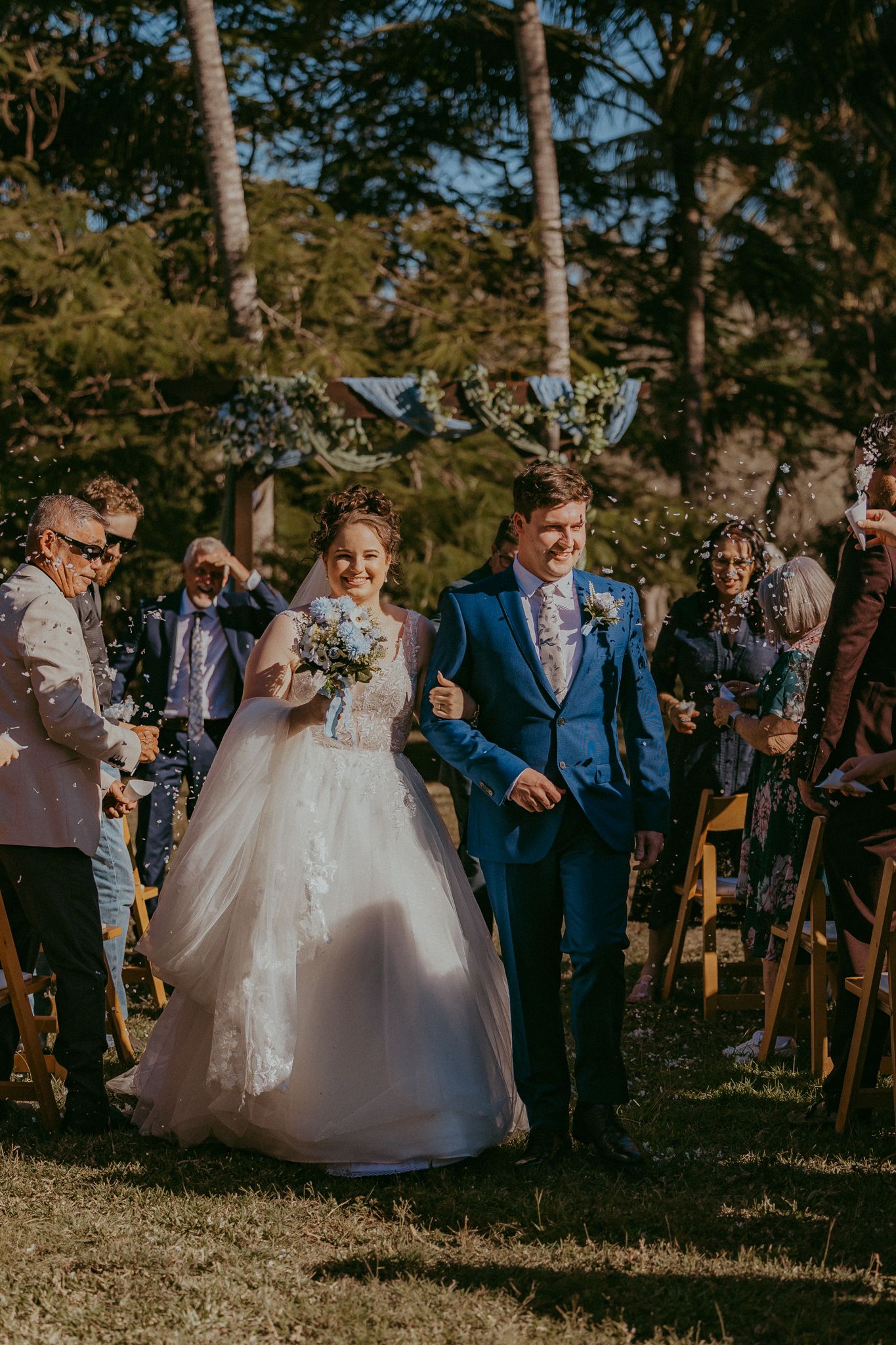 Newlyweds walk down the aisle as guests toss confetti at a sunny outdoor wedding ceremony.