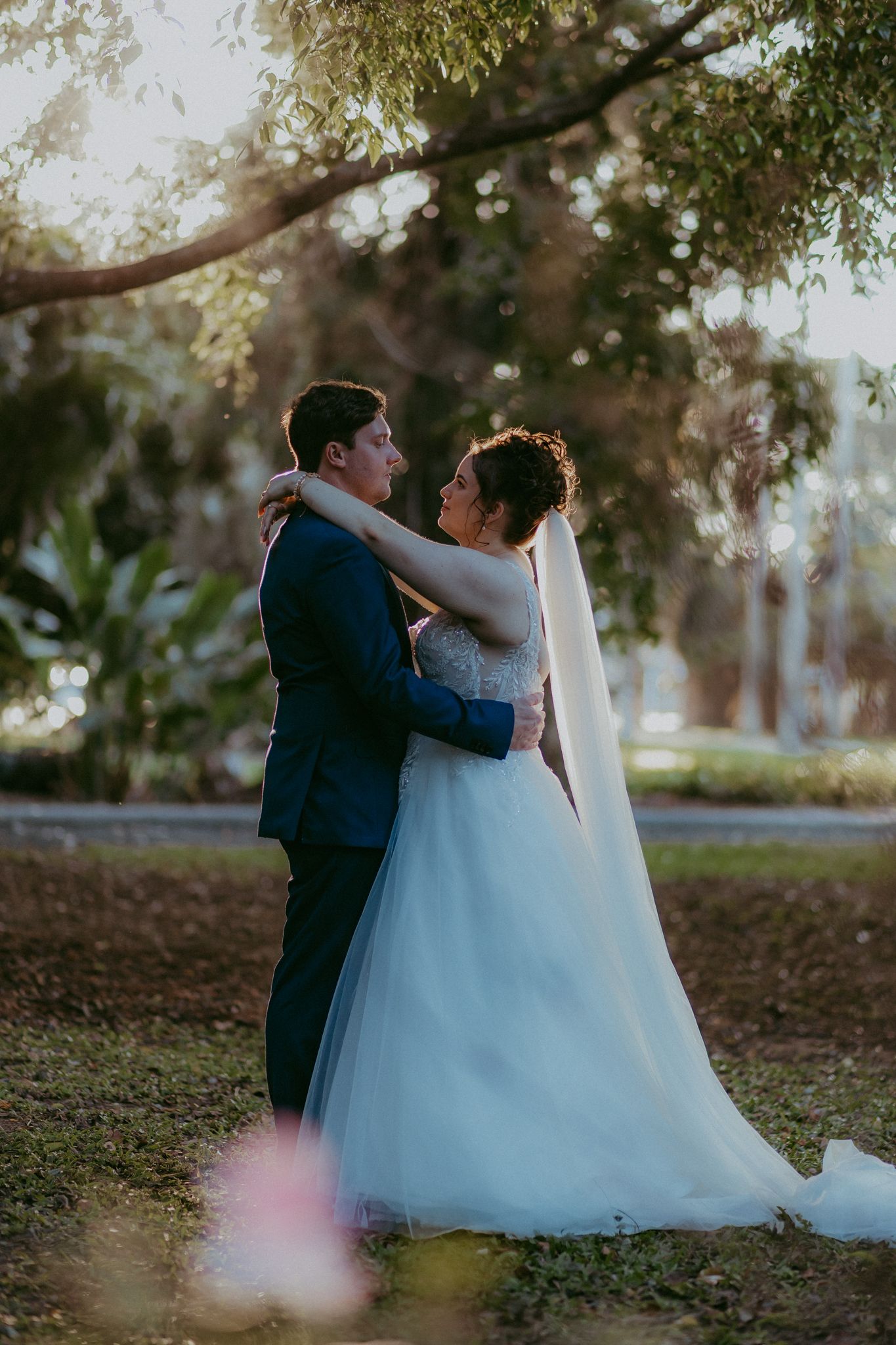 Bride and groom share a romantic embrace under a tree in golden hour light at an outdoor wedding.