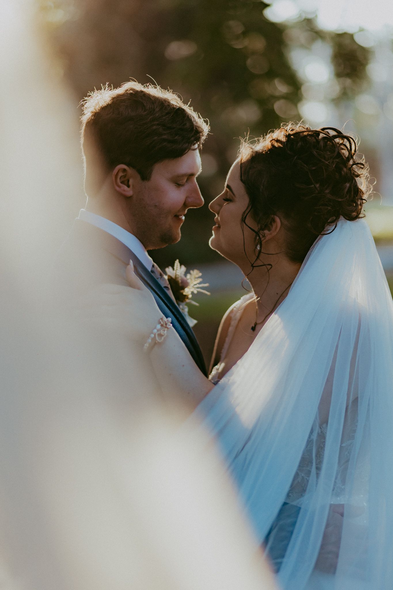 A bride and groom lean in for a kiss, framed by soft bokeh and warm golden light outdoors.