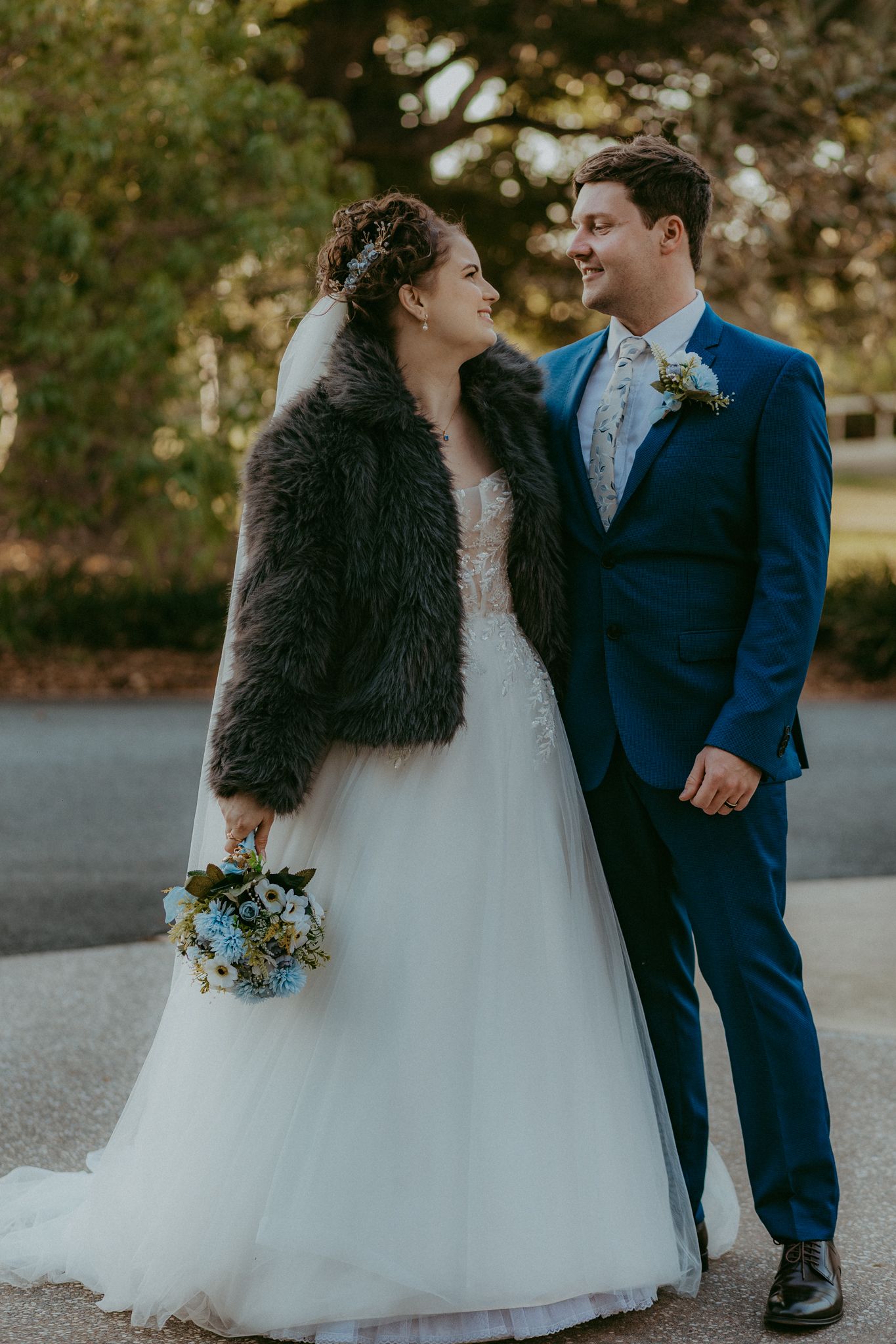 Bride in white gown and fur jacket holds blue bouquet while smiling at groom in blue suit.