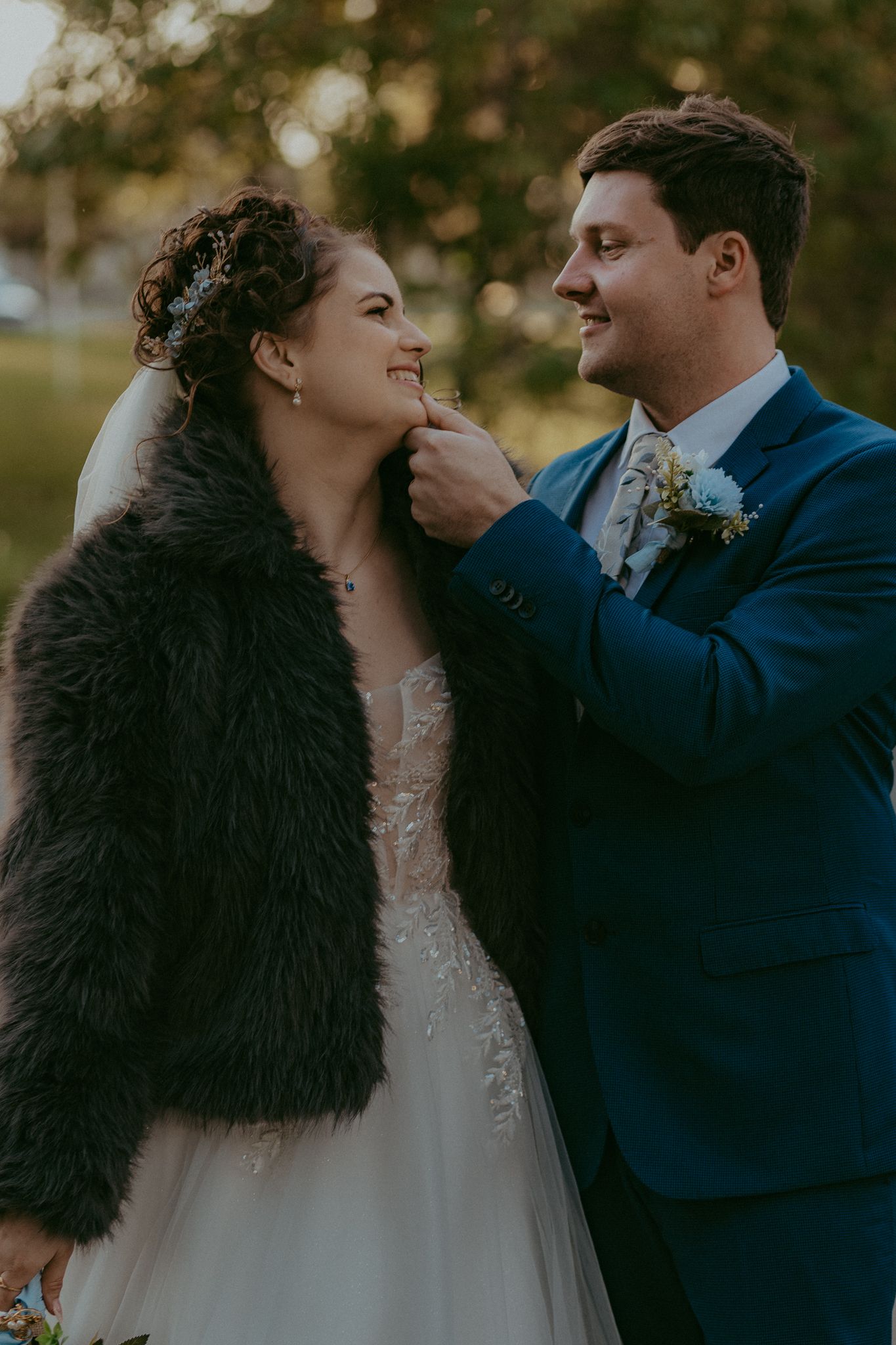 Groom in blue suit tenderly touches bride's face as she smiles in a fur jacket outdoors.