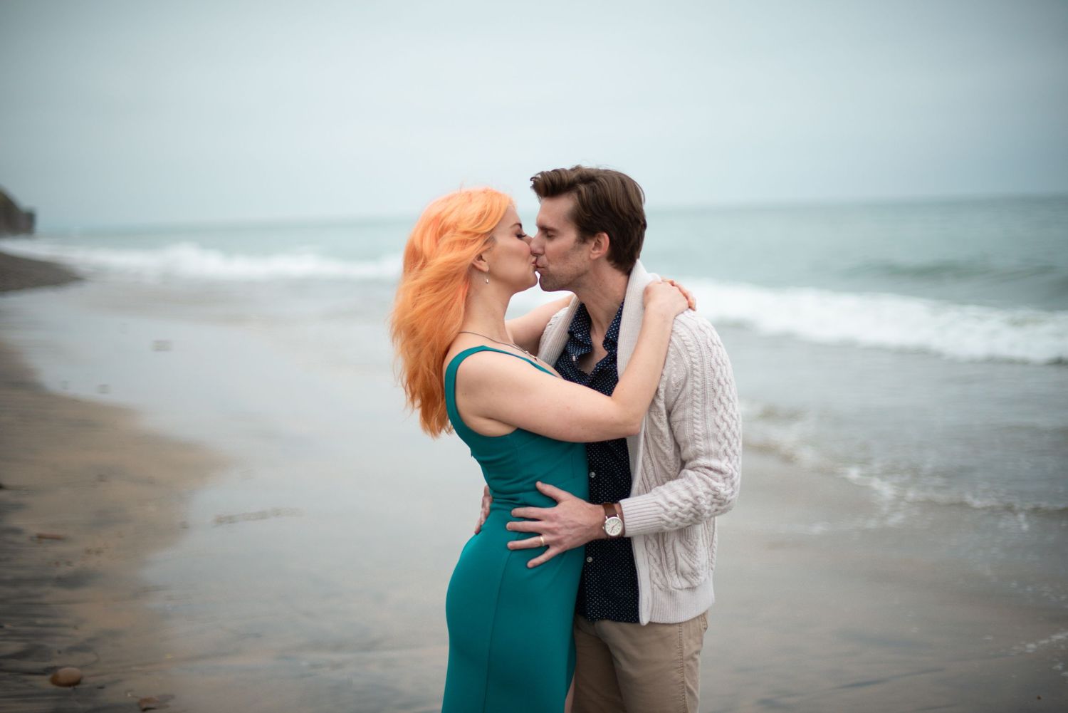 A couple embraces on a misty moonlight beach in Encinitas, with her vibrant red hair and teal dress contrasting against the grey shoreline.