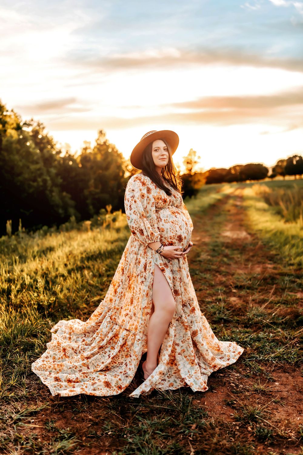 A floral dress flows elegantly in the golden sunset light along a rustic dirt path through grassy fields.