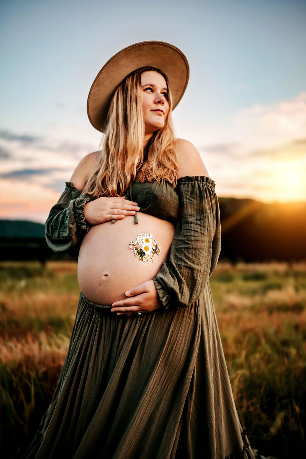 Pregnant woman in olive off-shoulder dress and tan hat cradles bare belly with daisy at golden hour sunset.