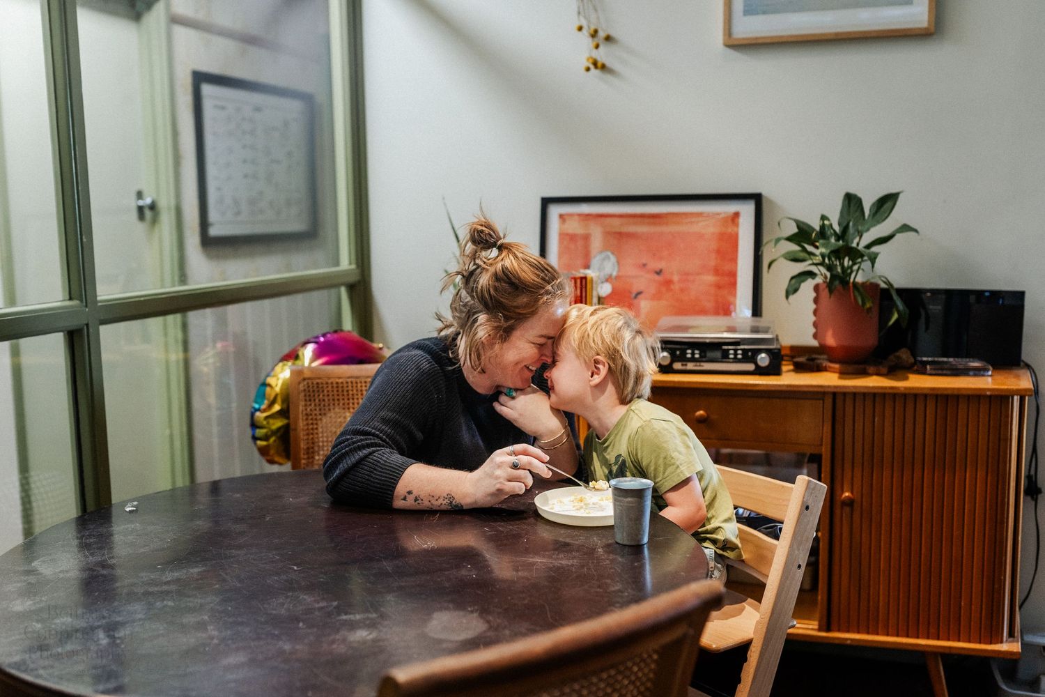 A mother and her three-year-old son place their foreheads against each other, while sitting at the dining table eating morning tea.