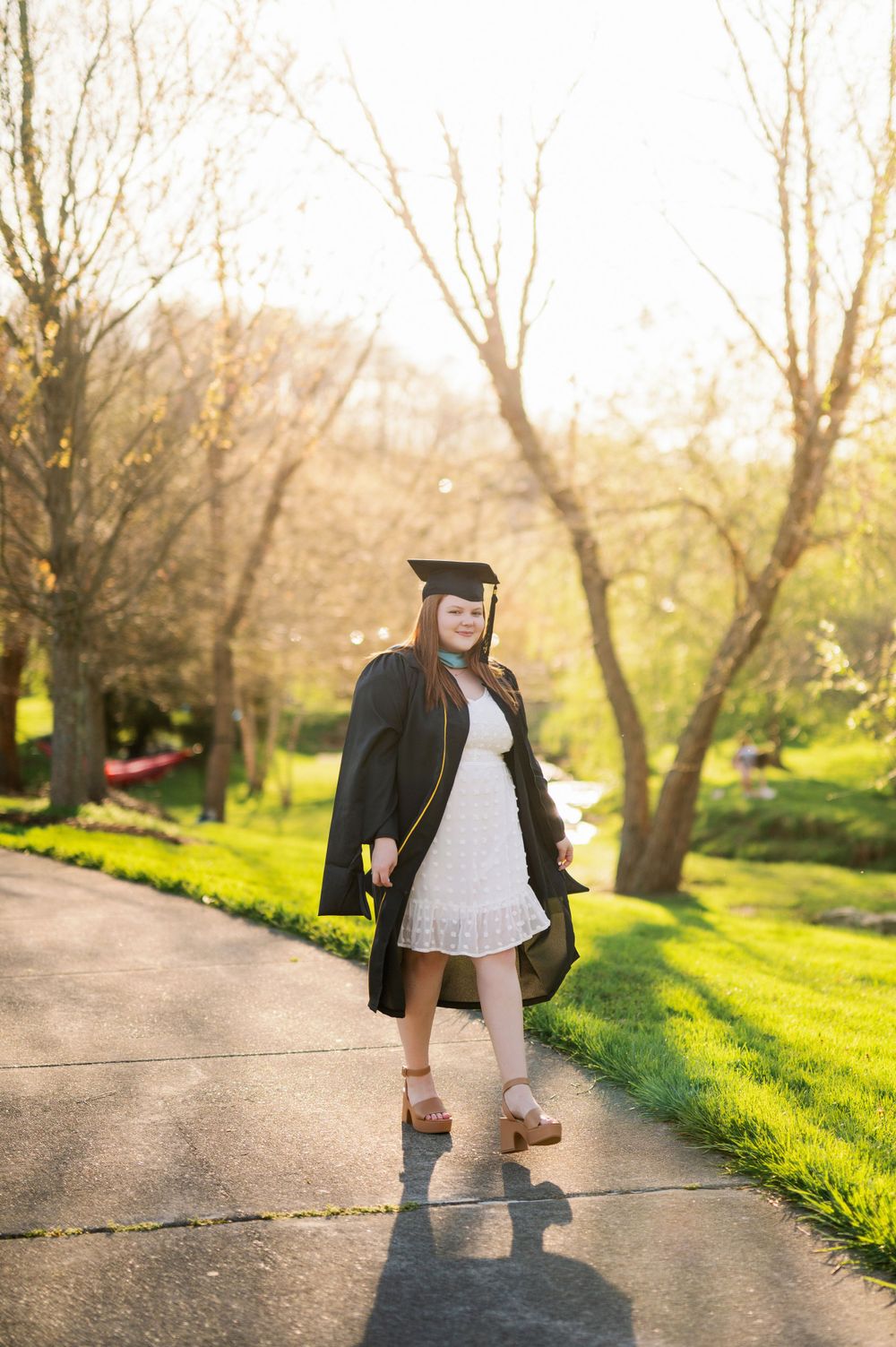 Graduate in cap and gown walking on sunny path during spring.