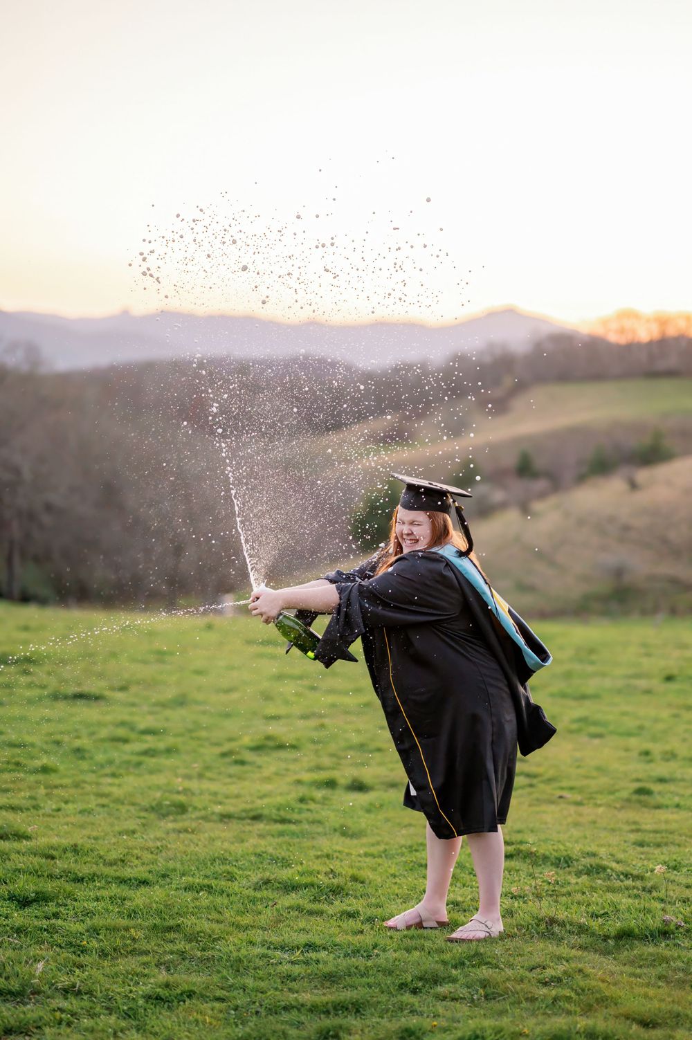 Graduate in cap and gown spraying champagne in celebration on hilltop.