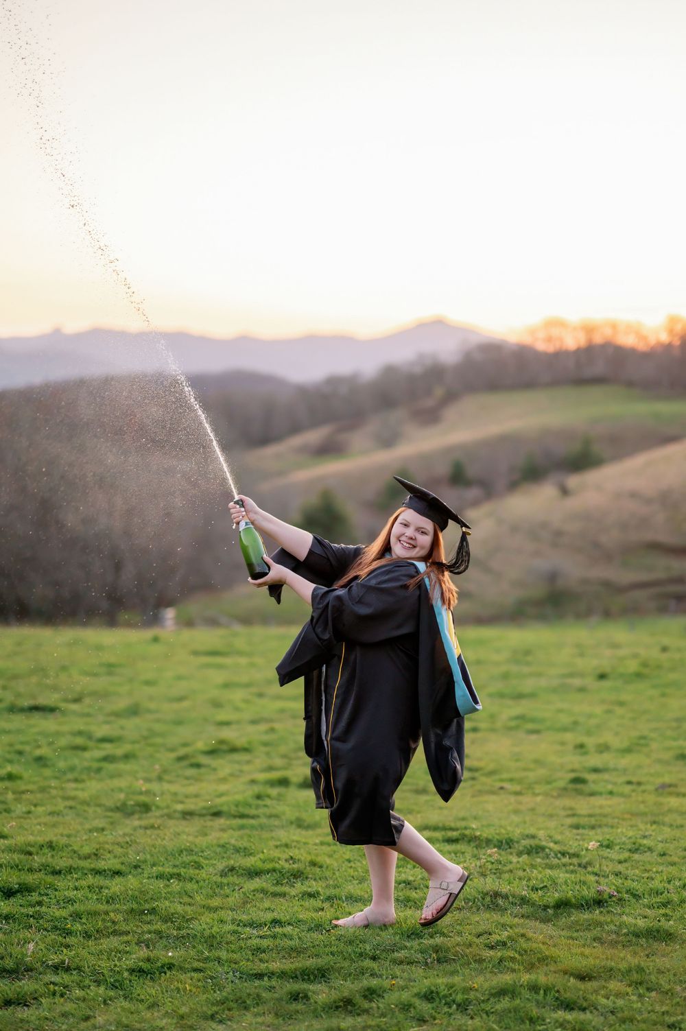 Graduate in black cap and gown celebrates by spraying champagne on a scenic hilltop at sunset.
