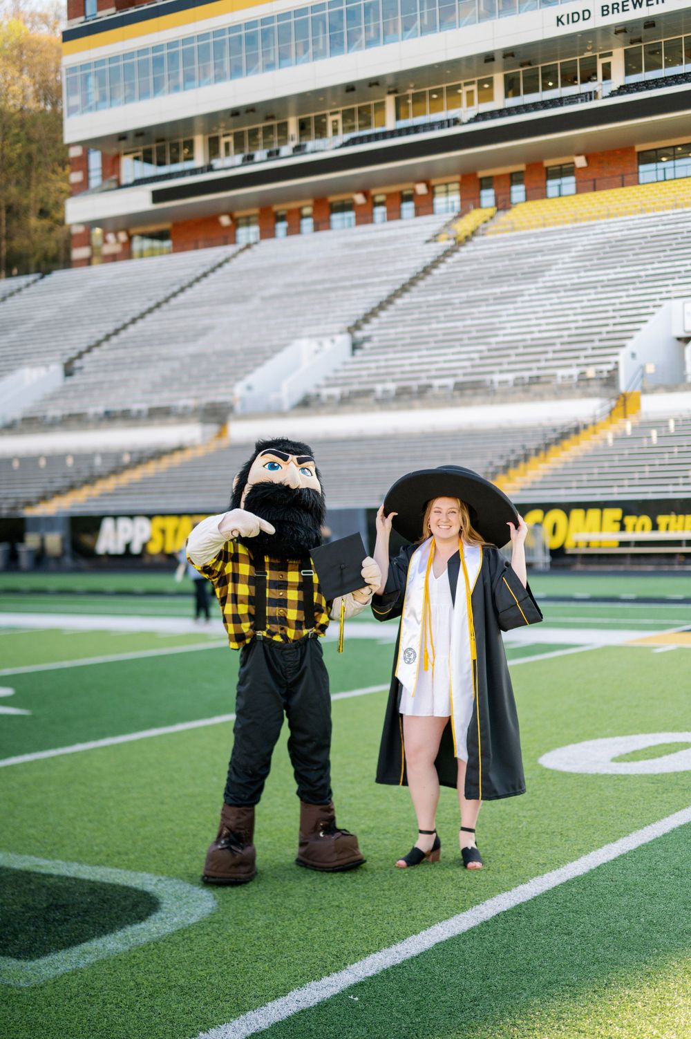 Graduate poses with school mascot on football field in front of stadium bleachers.