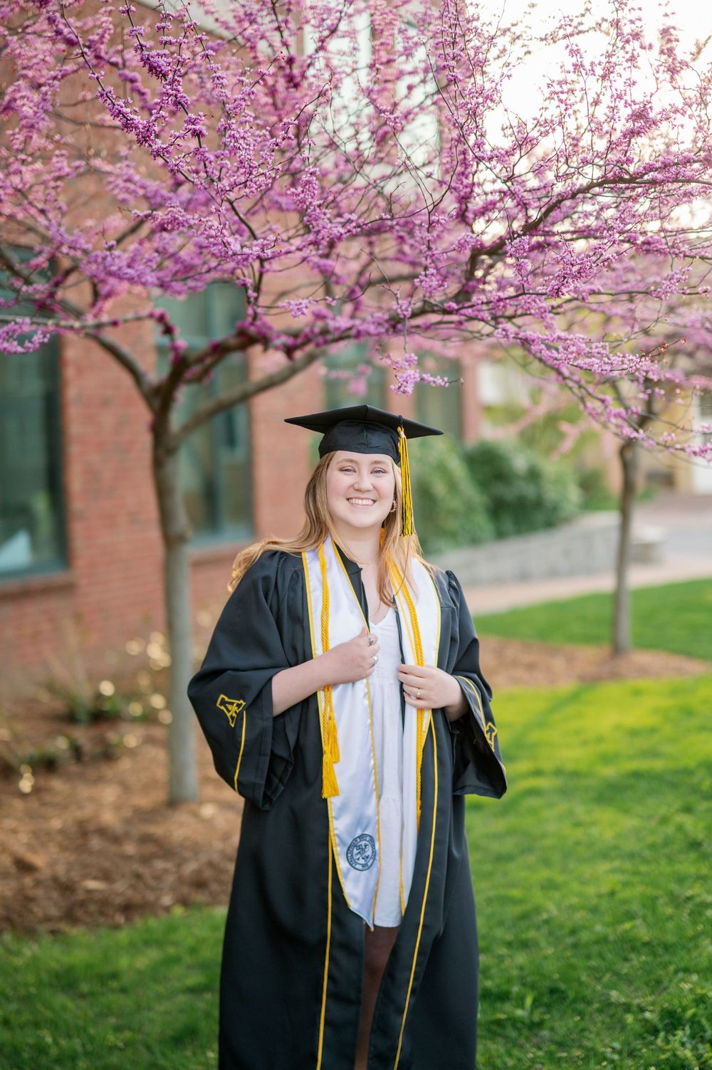 Graduate in black cap and gown poses under blooming cherry blossom trees on campus.