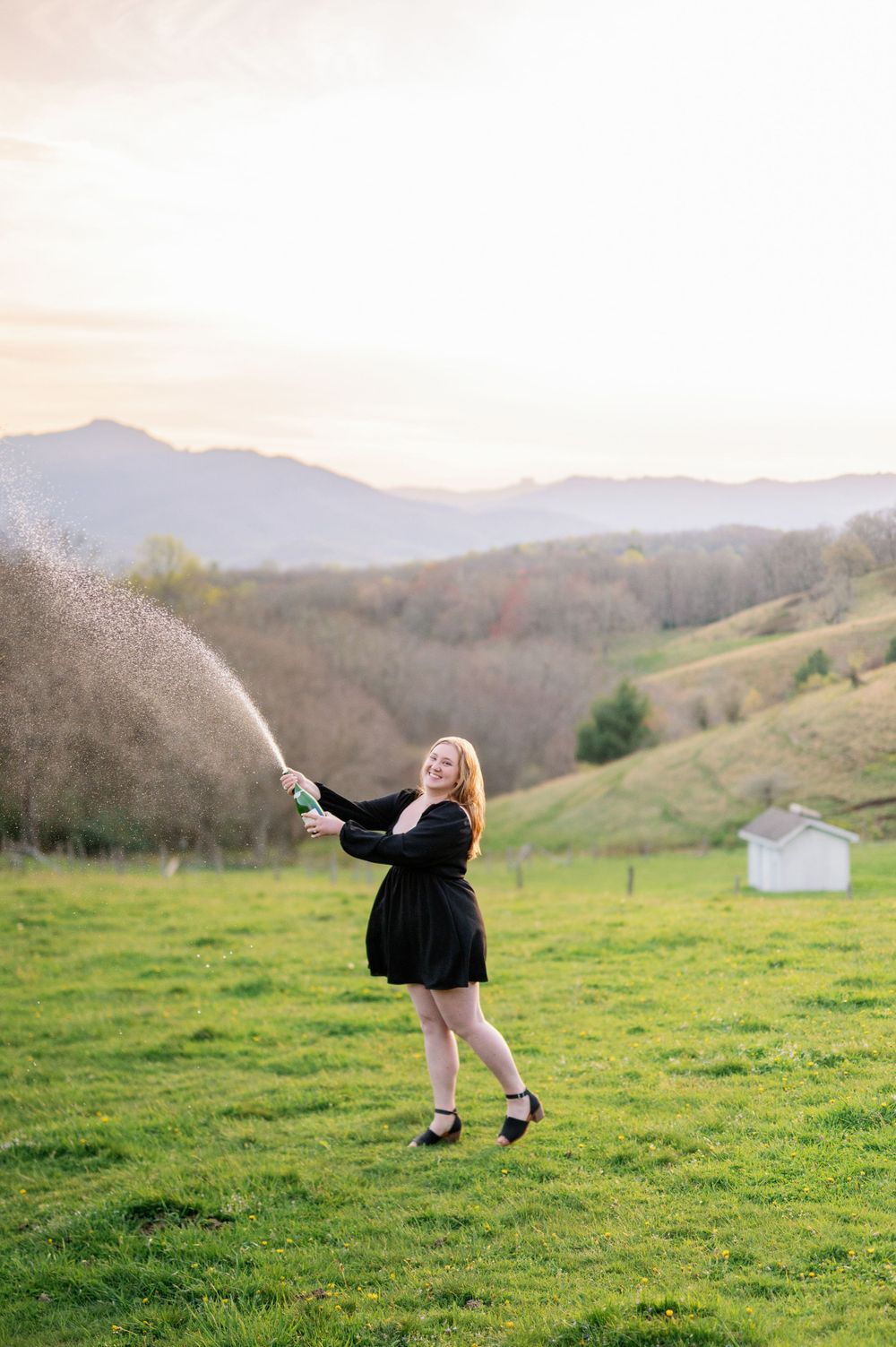 Graduate in black dress celebrates with champagne spray in rolling mountain landscape.