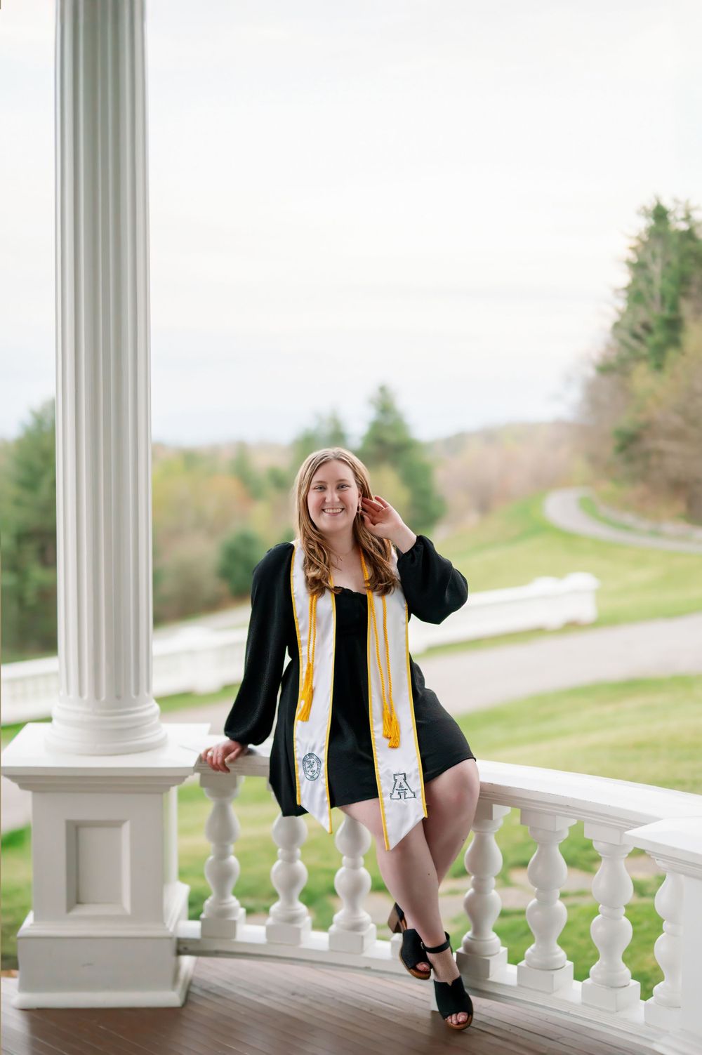 Graduate in academic regalia poses on white columned porch with mountain views.