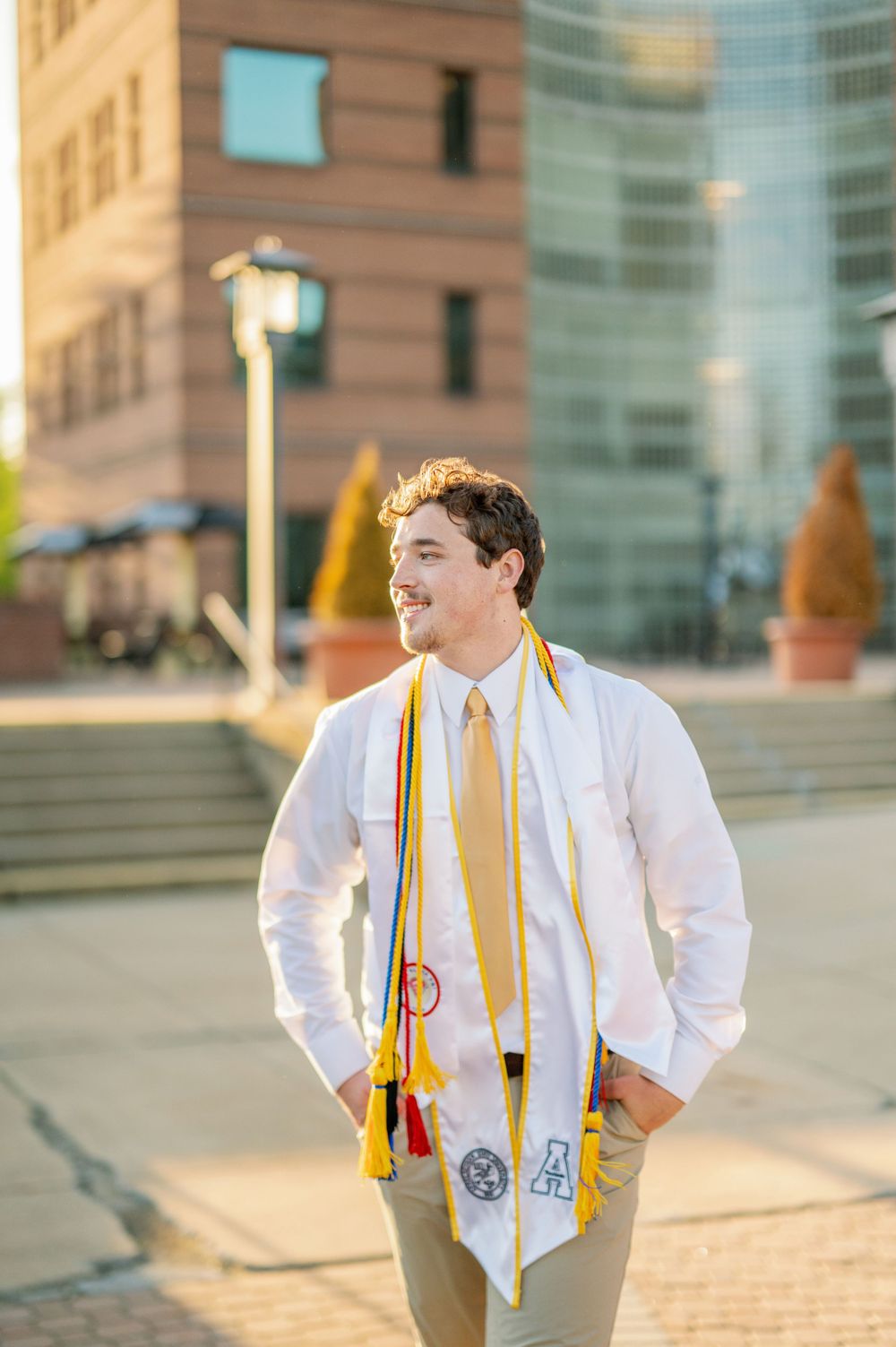 Graduate in white shirt and gold stole stands outside modern university building.
