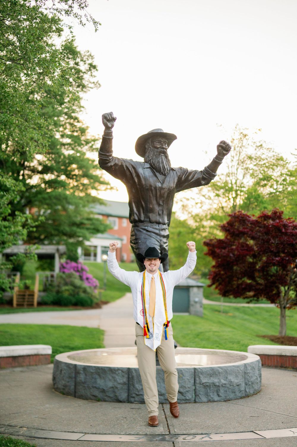Graduate poses with triumphant statue on university campus during graduation.
