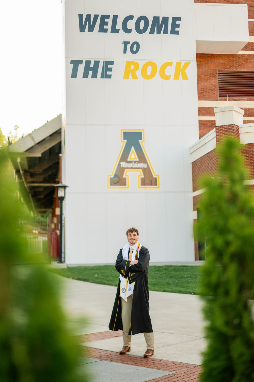 Graduate stands before Welcome to The Rock sign at Appalachian State University.