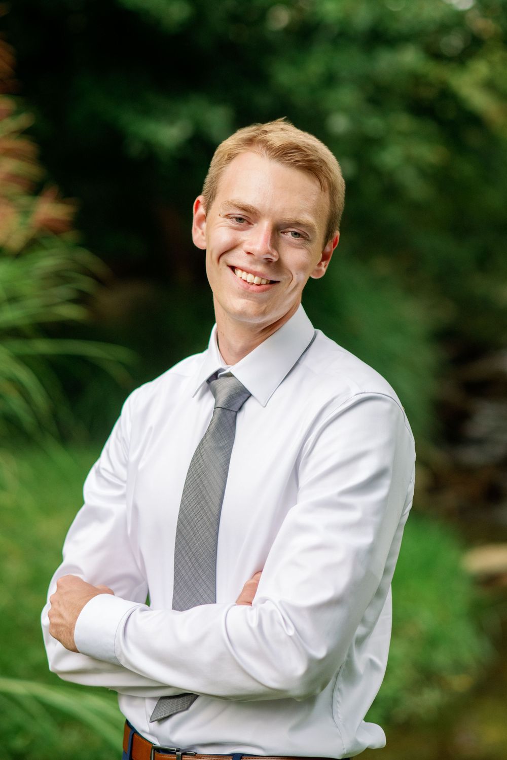 Smiling young man in white shirt and gray tie with arms crossed outdoors among green trees.