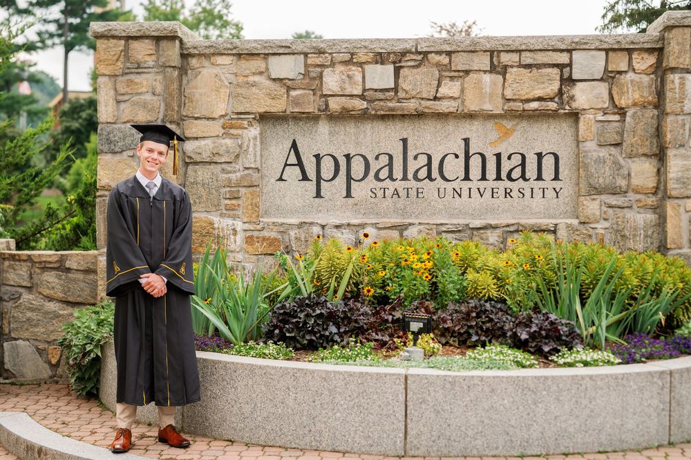 Graduate in cap and gown poses proudly in front of Appalachian State University stone entrance sign.