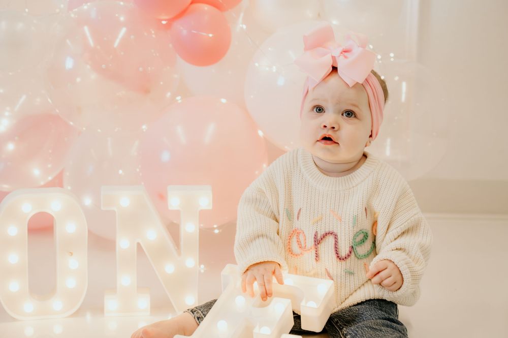 Baby with pink balloons and decorations in a soft, dreamy setting.