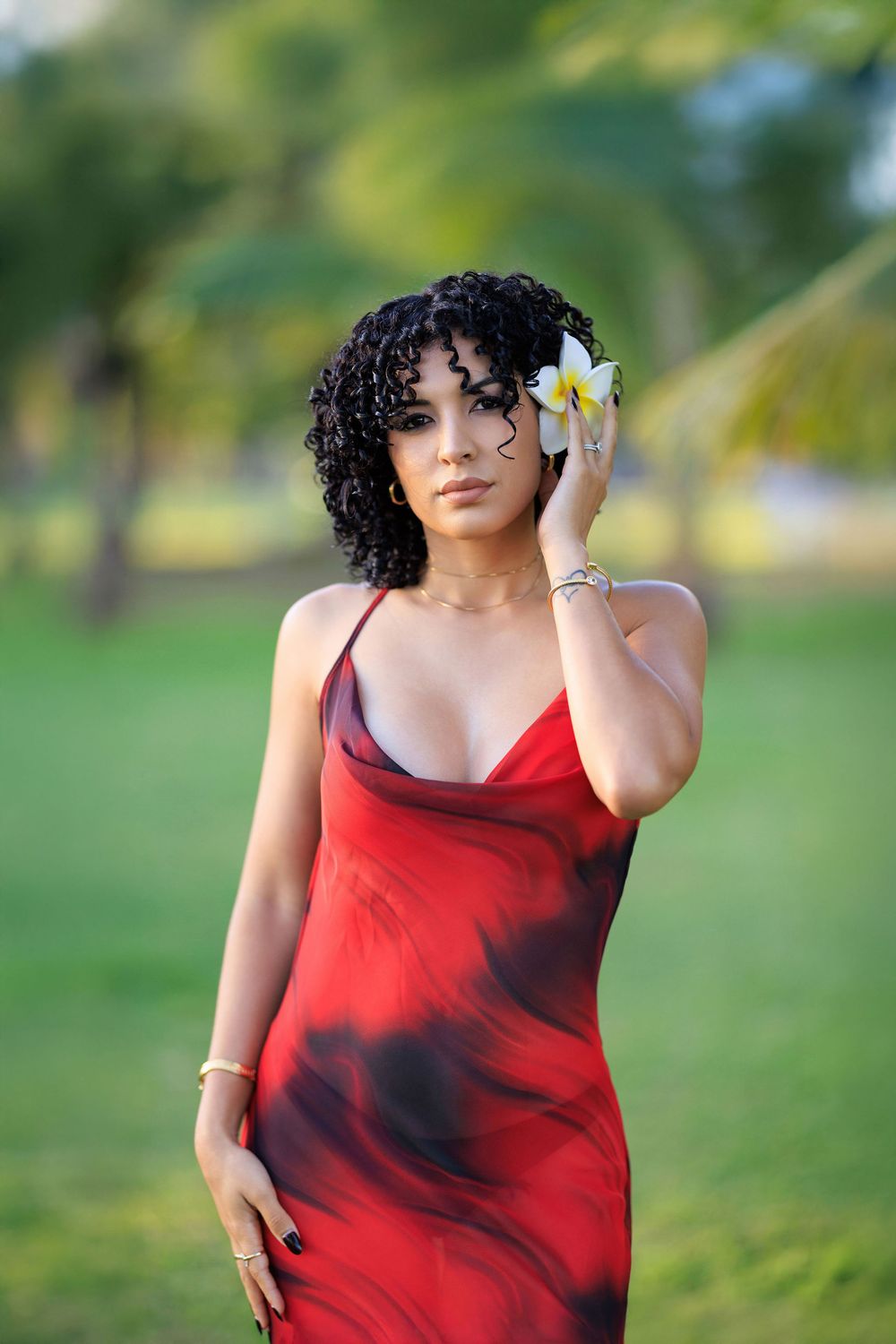 Woman in red tie-dye slip dress with curly hair and white flower accessory posing outdoors near palm trees.
