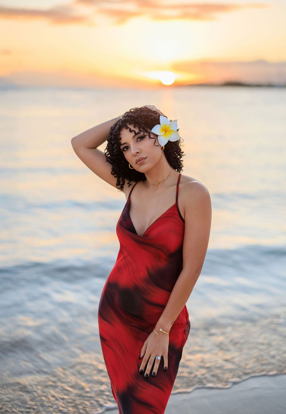 Woman in red tie-dye dress with plumeria flower in hair poses on beach at sunset.