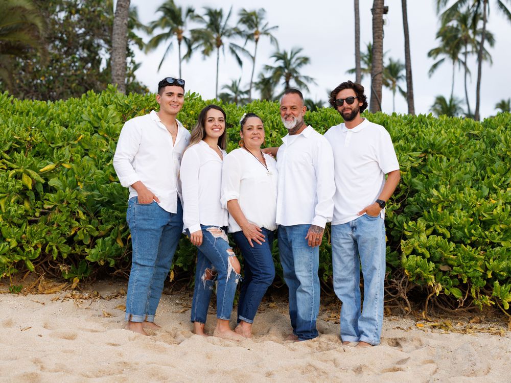 Family of five in matching white shirts and jeans posing on sandy beach with tropical palm trees and green hedges.