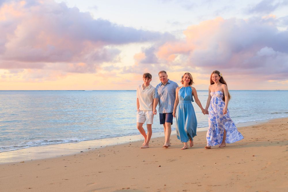 Family of four walking hand-in-hand on a sandy beach at sunset with colorful pink and blue sky.