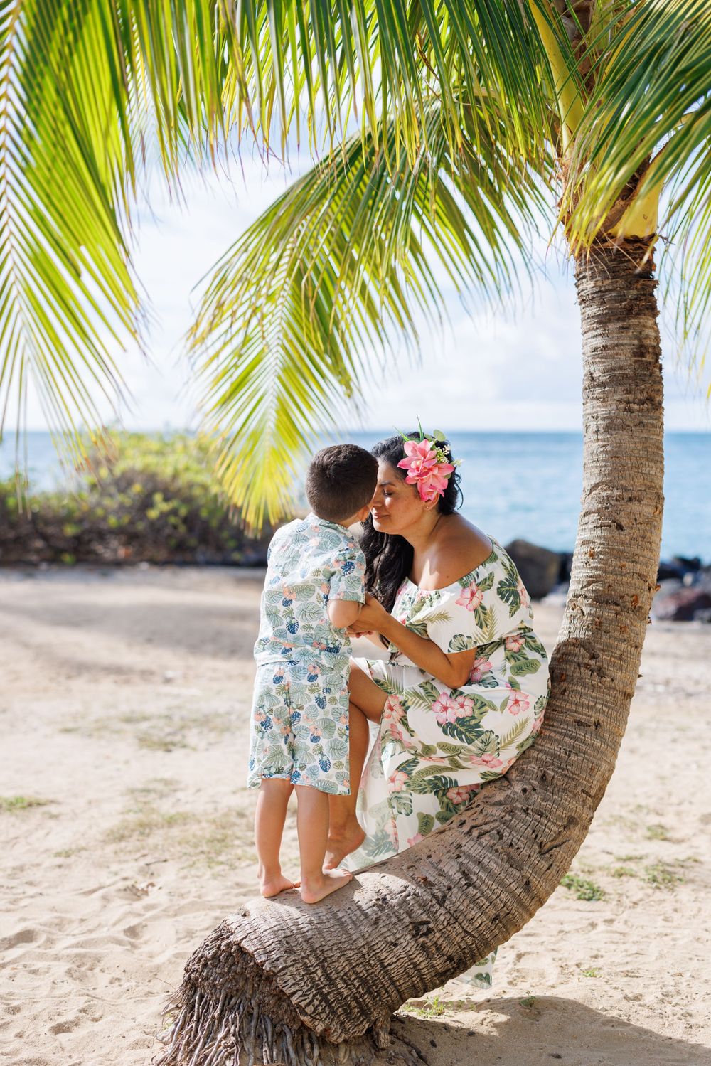 Pregnant woman and child kissing on a leaning palm tree on a tropical beach in matching floral outfits.