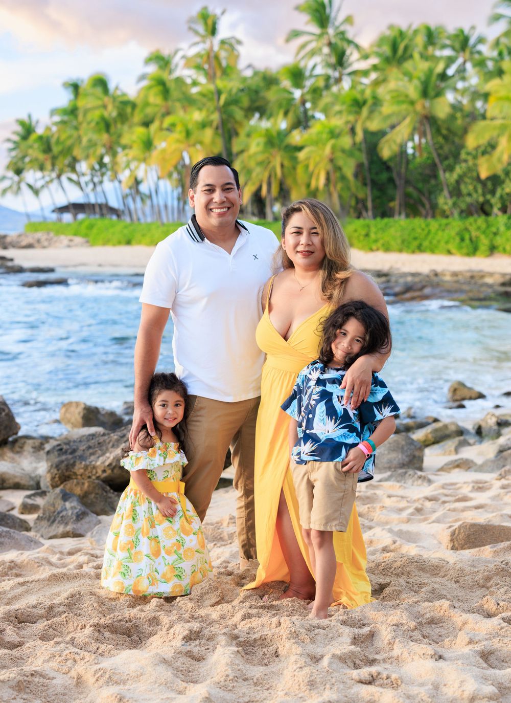 Family of four posing on a sandy beach with palm trees, wearing bright summer outfits in a tropical setting.