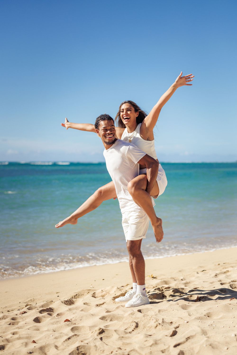 Happy couple in white outfits celebrating on a sunny tropical beach, man giving woman a piggyback ride with ocean backdrop.