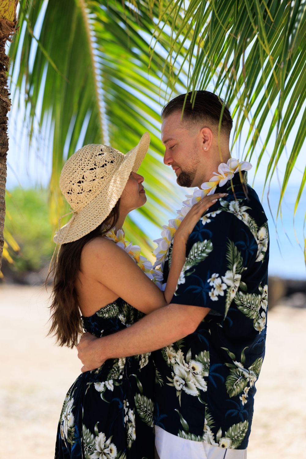 Couple in matching Hawaiian floral outfits embracing under a palm tree on a tropical beach.