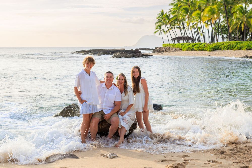Family of four posing on a sandy Hawaiian beach at sunset, wearing white outfits with ocean waves and palm trees.