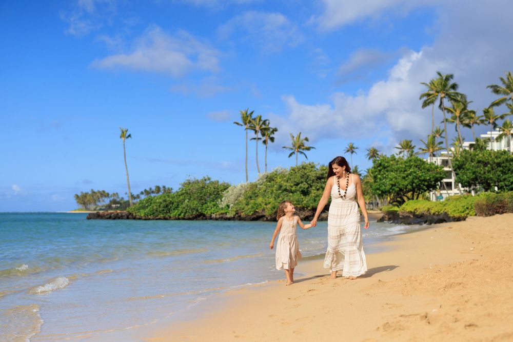 Mother and daughter walking hand in hand on a sunny Hawaiian beach with palm trees and blue ocean.