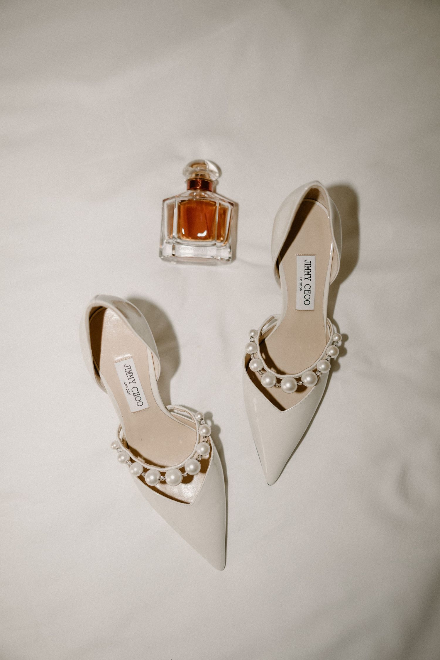 Wedding accessories including white stiletto heels, perfume bottle, and floral details arranged on white fabric.