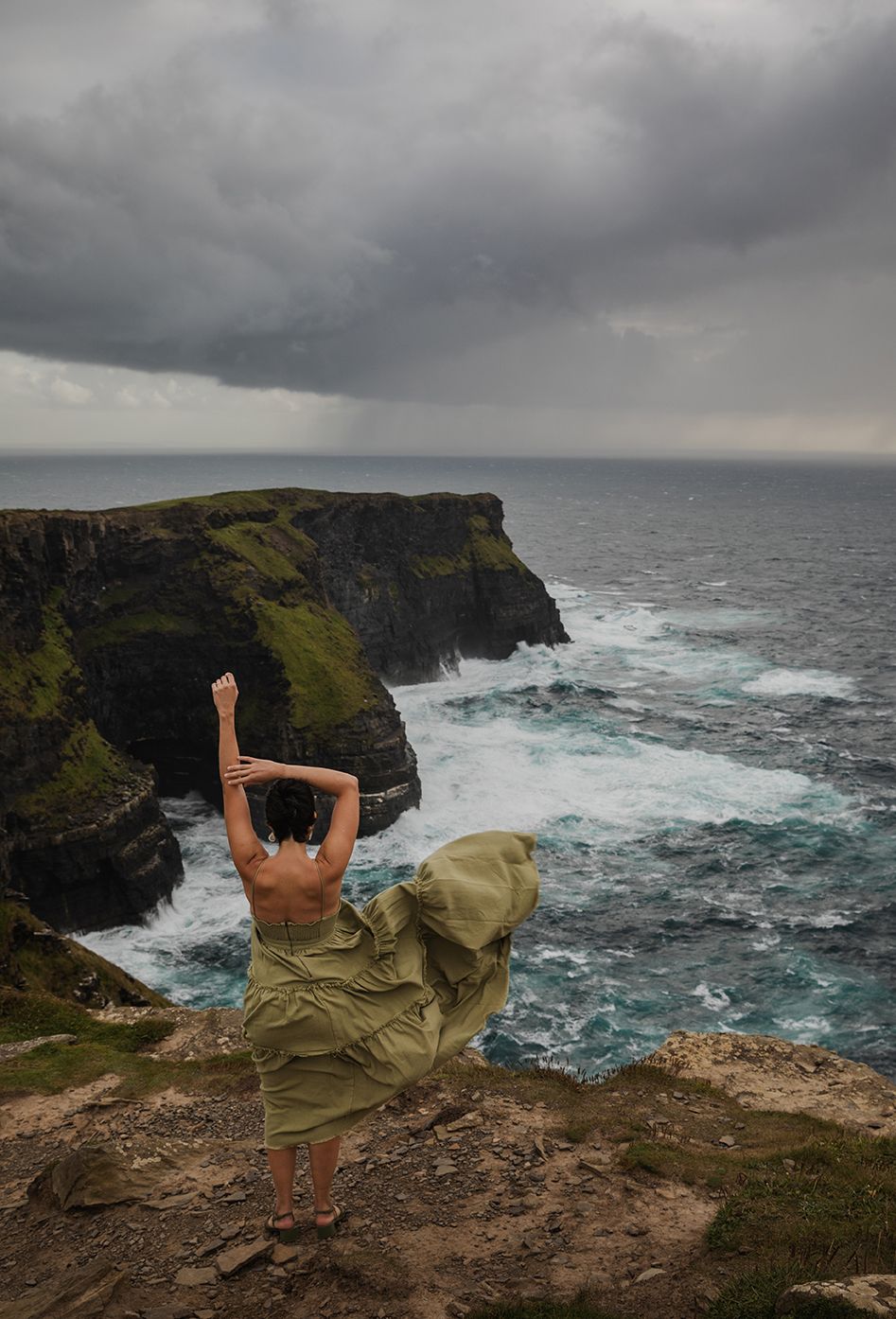 woman in a long green dress is standing at the edge of Cliffs of Moher during her adventurous portrait photo session