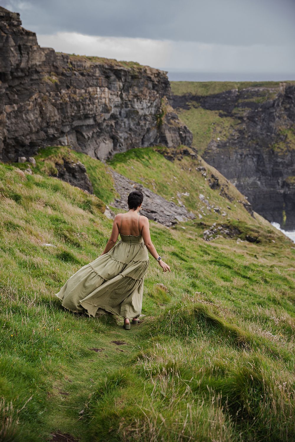 woman in a long green dress is walking along the Cliffs of Moher during her adventurous portrait photo session