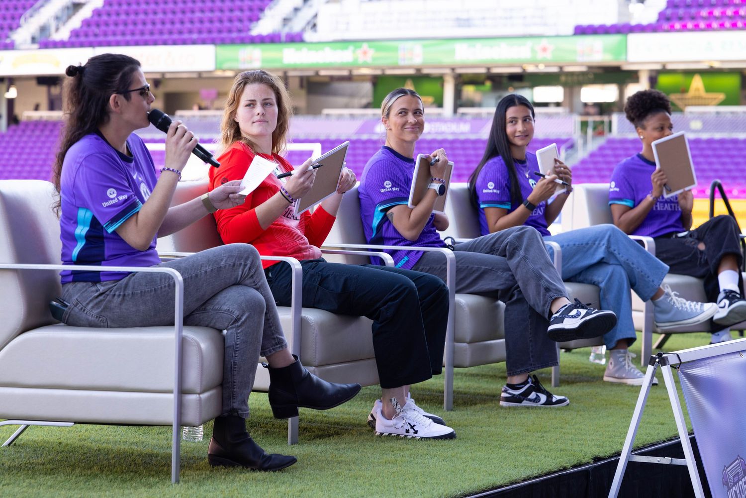 People seated on benches at a stadium with purple seating in the background.