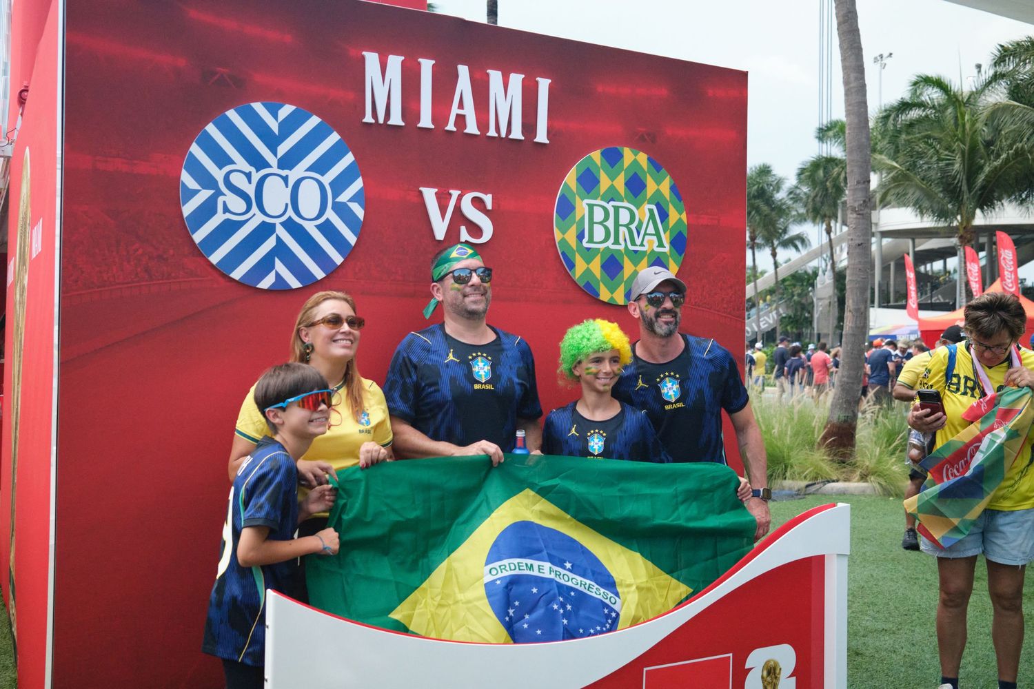 Brazil fans pose with flag at Miami SCO vs BRA match venue, wearing green jerseys and colorful wigs.