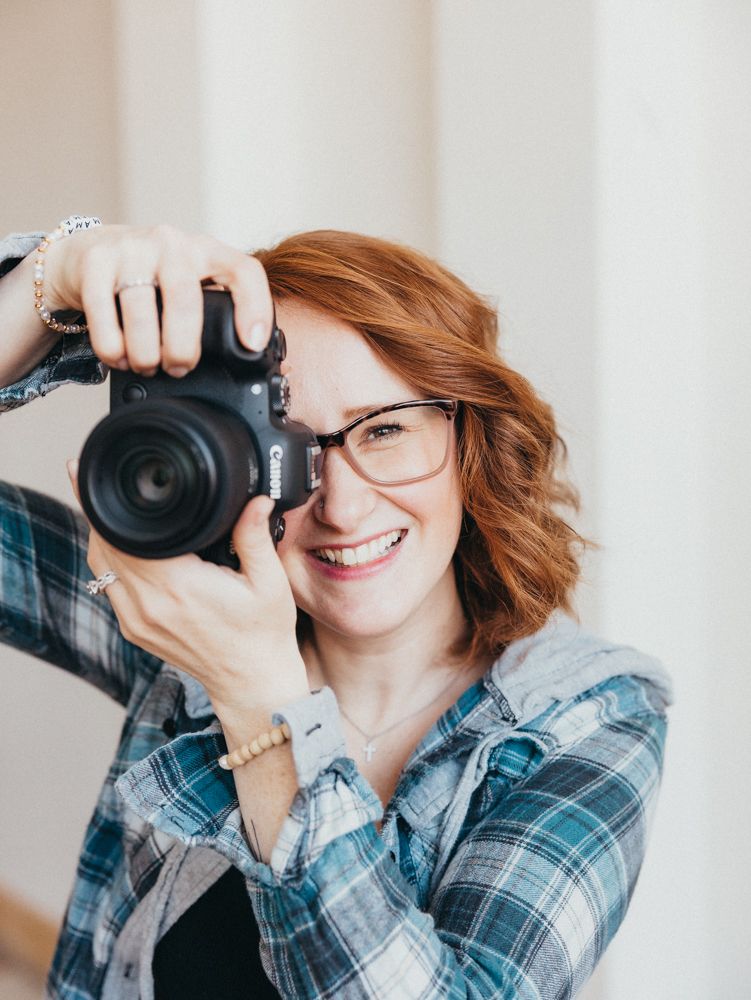 Photographer in brown dress holding camera while sitting on bed.