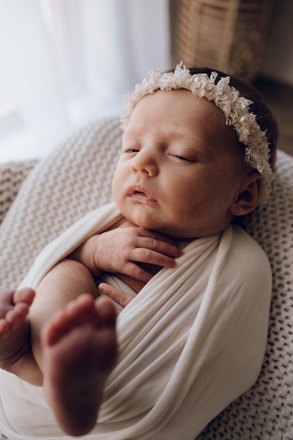 Close-up of sleeping newborn in white wrap with lace headband showing tiny feet.