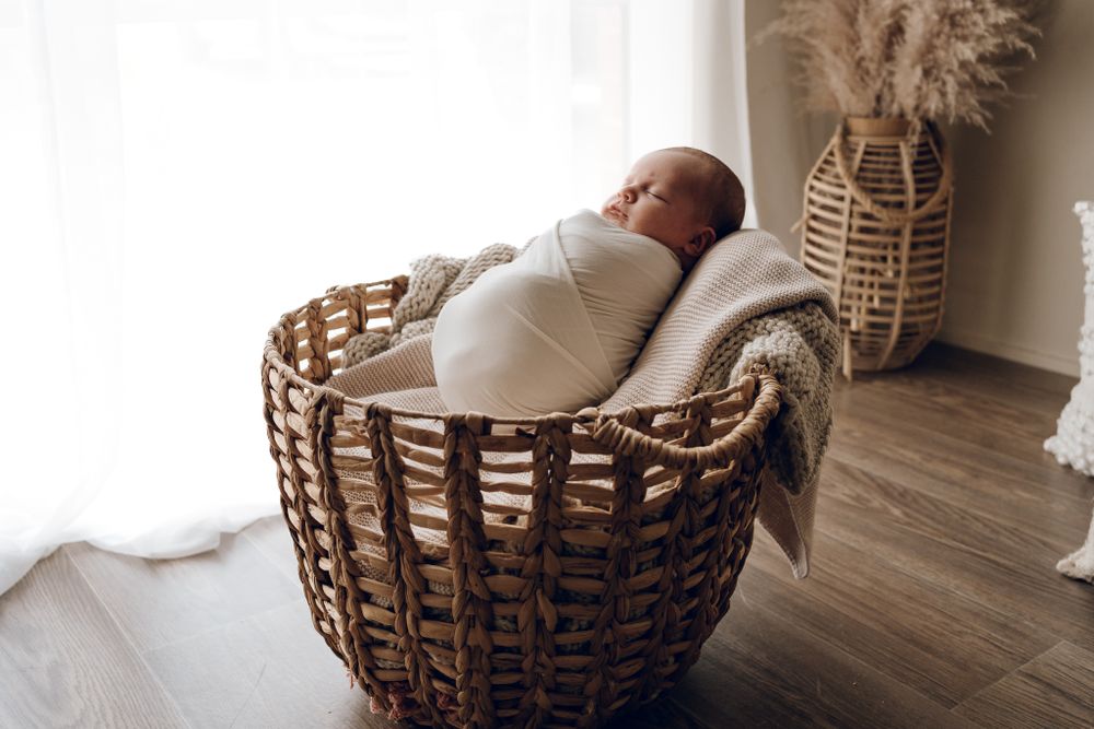 Sleeping newborn swaddled in white lies peacefully in woven basket with pampas grass decor.