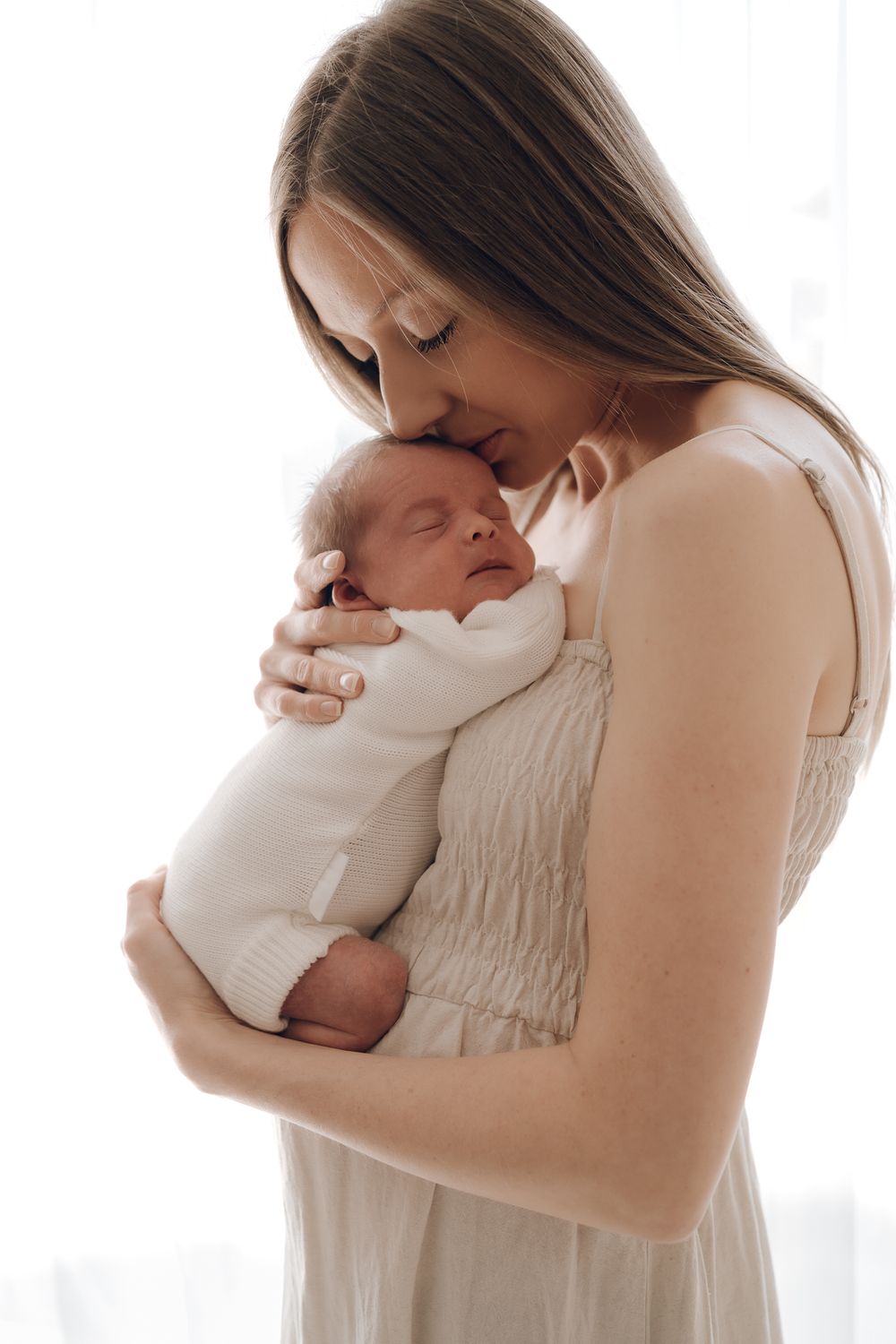 Mother in white dress holds newborn baby in a bright, minimalist setting.
