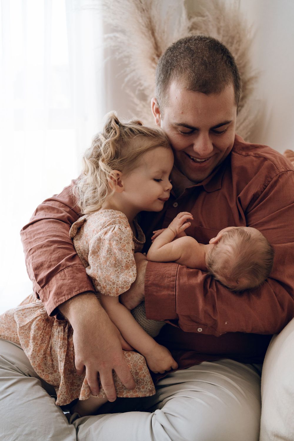 Parent in rust-colored jacket snuggles with two small children on a couch in an intimate family moment.