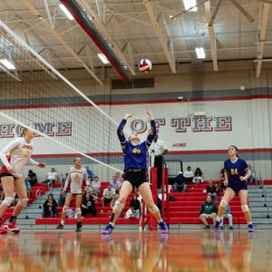 Indoor volleyball match with players from opposing teams jumping at the net during an intense play in a school gymnasium.