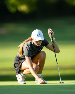 Female golfer in Nike cap crouches to read the green while holding putter on sunny golf course.