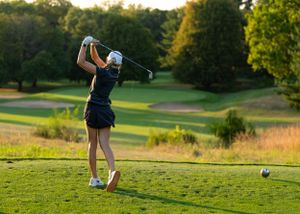 A golfer in a navy dress swings a club at sunset on a scenic golf course with rolling hills and lush trees.