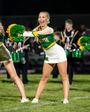 A cheerleader in green and white uniform performs an energetic dance routine on a sports field at night.