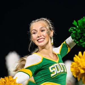 A cheerleader in green and yellow uniform performs during a game while holding bright yellow pom poms.