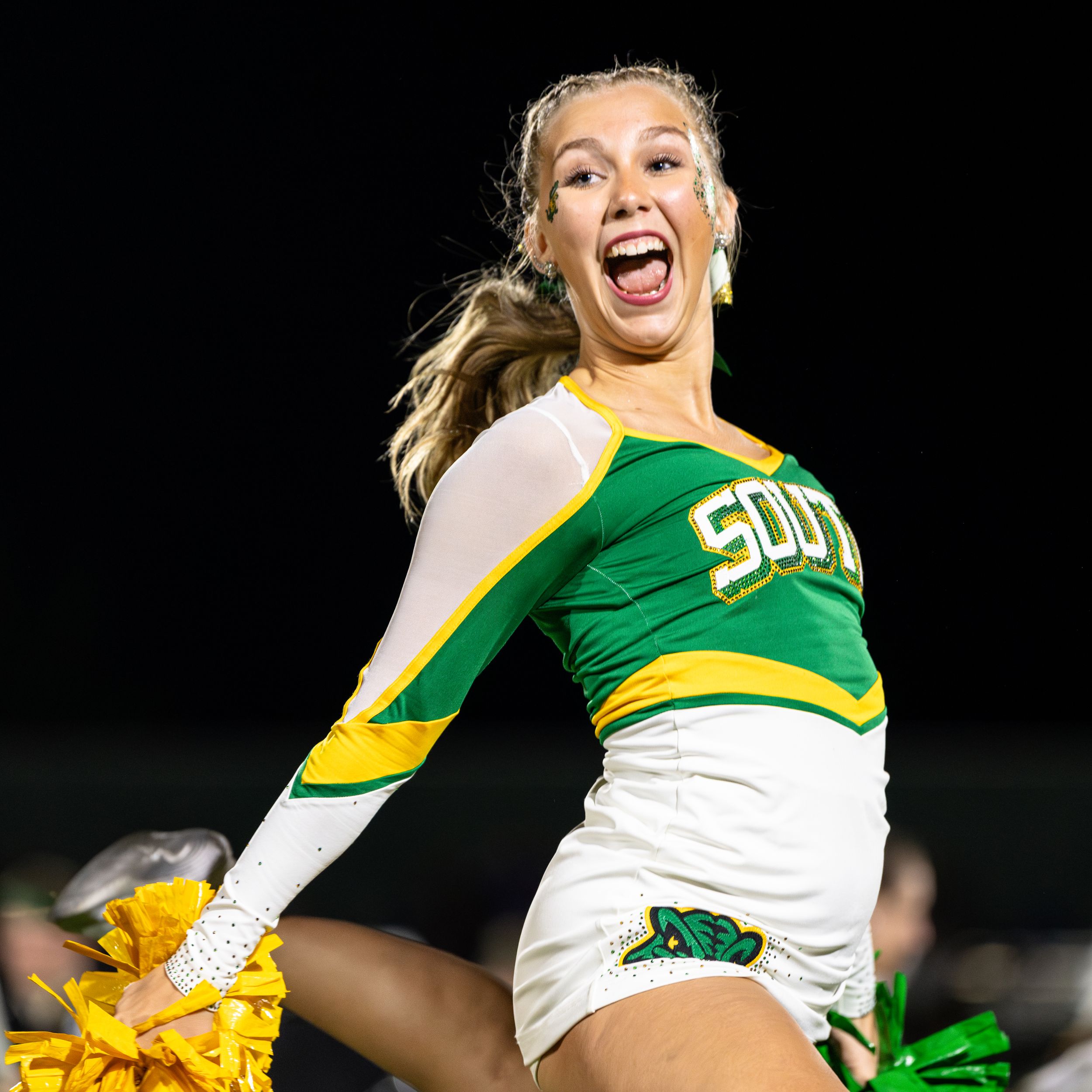 Cheerleader in green and white uniform performs energetic routine with pom poms against dark background.